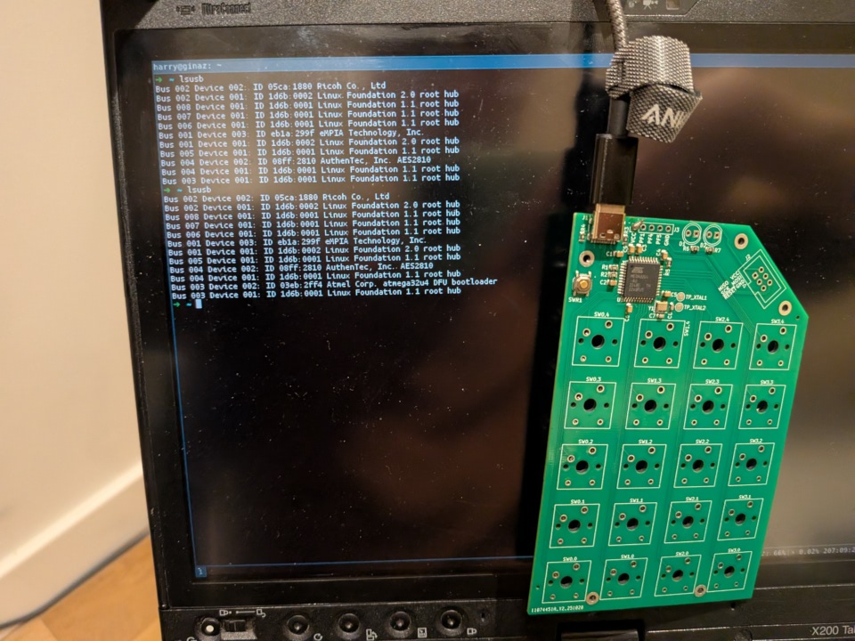 Photo of a TwentyPad PCB dangling by its USB connection in front of the screen of a vintage ThinkPad, which is showing the output of lsusb in a terminal. Among the entries is one for "Atmel Corp. atmega32u4 DFU bootloader".